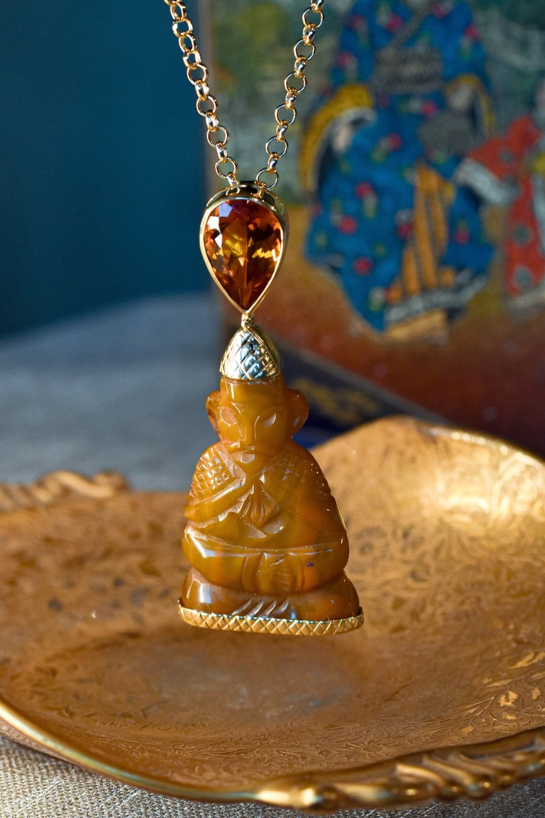 Gold chain necklace with carved amber Buddha pendant and amber teardrop top on ornate gold dish