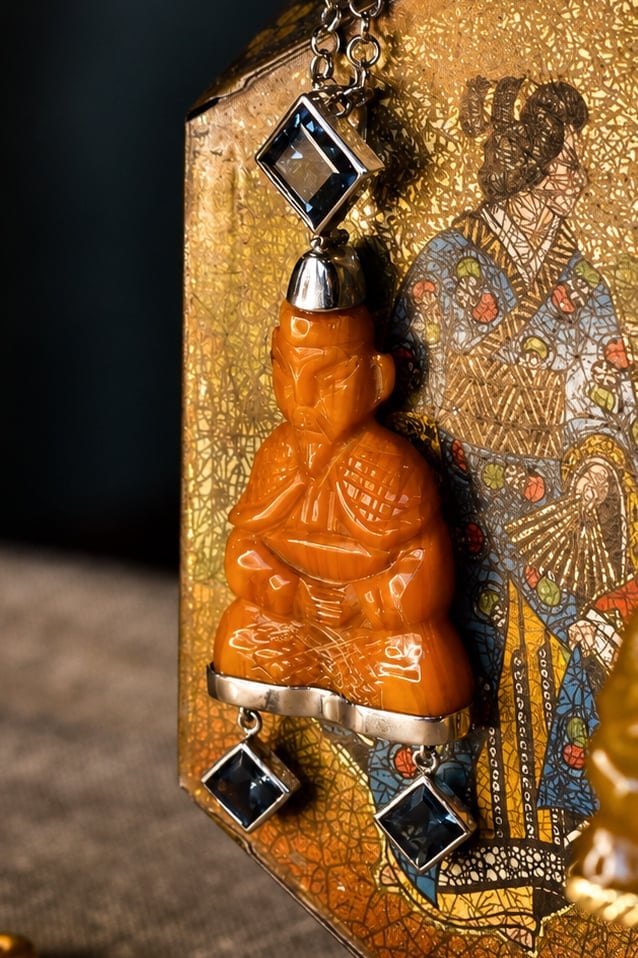 Orange carved Buddha pendant with black square stone settings, attached to a decorative gold-patterned box