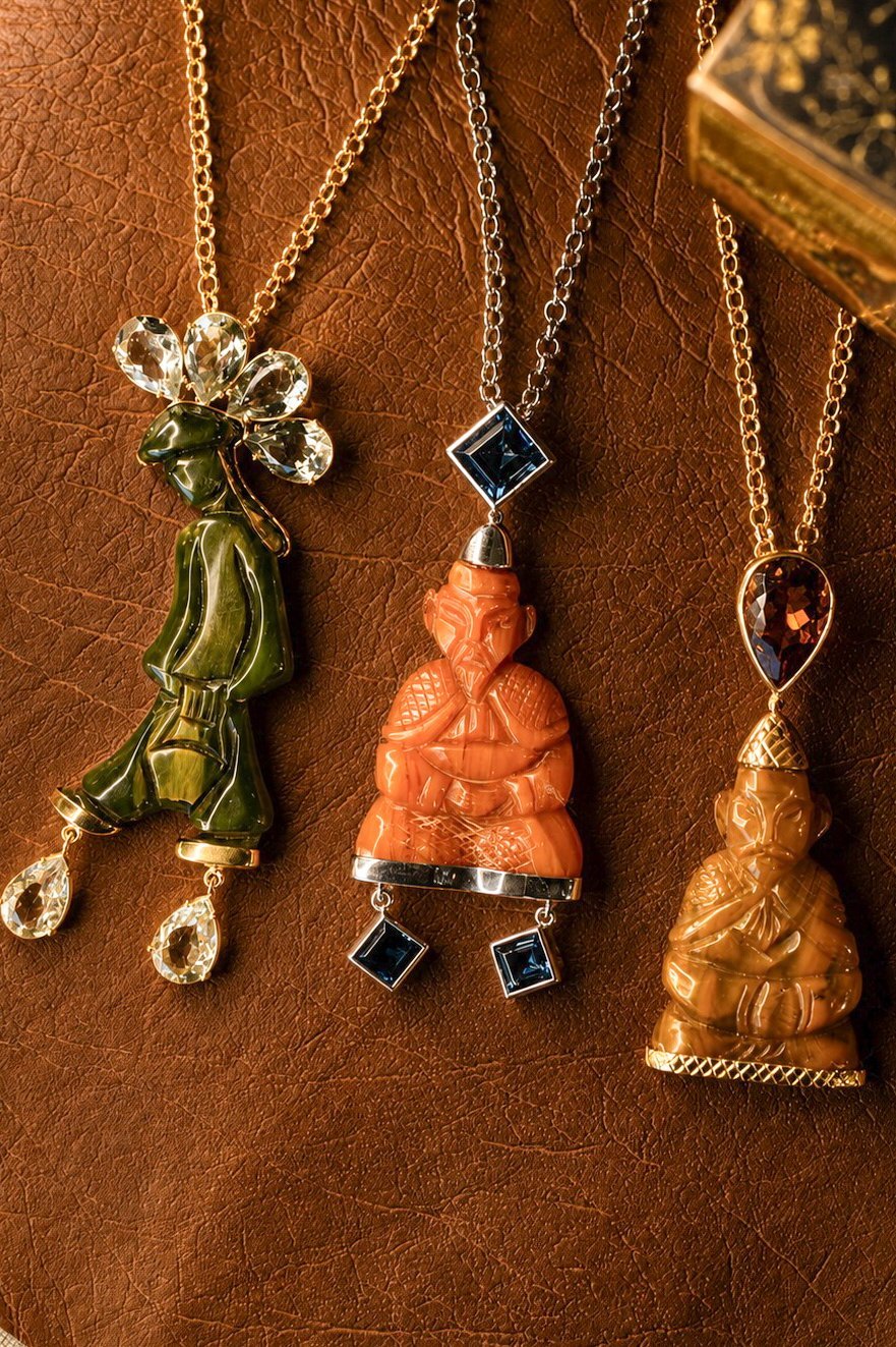 Three decorative pendant necklaces on gold chains displayed on brown leather: a green enamel pea pod, an orange carved Buddha with black stones, and a golden Buddha with a red gemstone