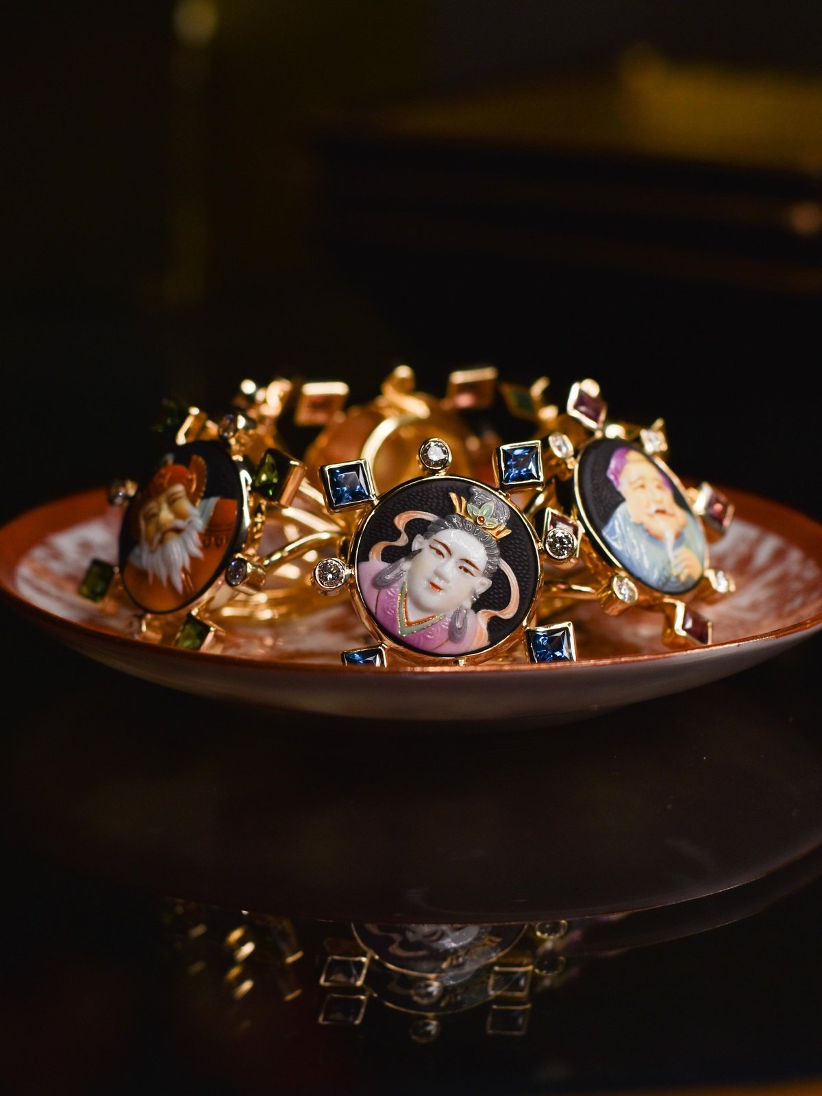Gold charm bracelet with portrait medallions and gemstones displayed on a copper dish against a dark background